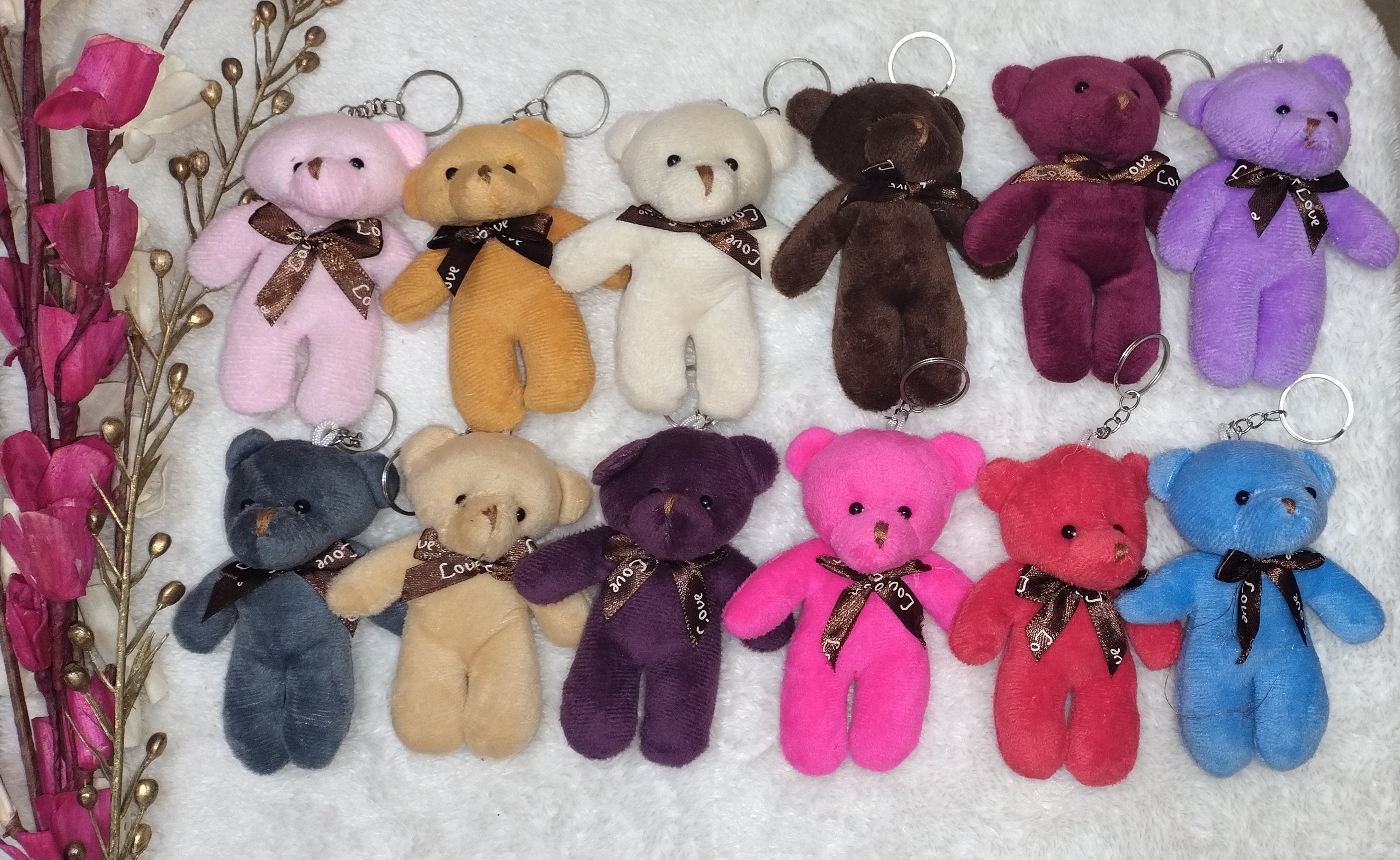 Colorful teddy bear keychains arranged on a white towel with a wooden surface and pink flowers in the background.