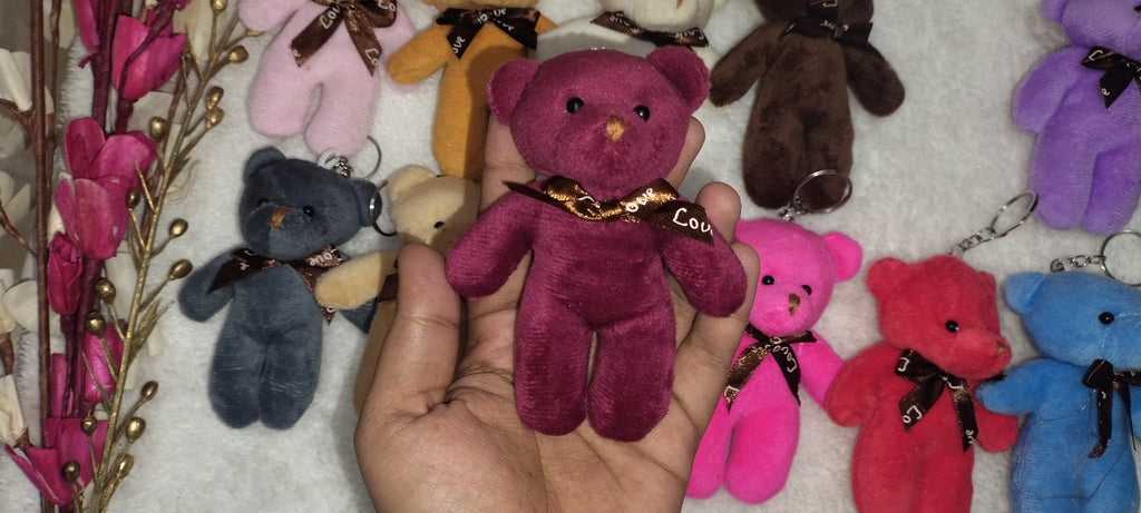 Hand holding a small purple teddy bear keychain with other colorful teddy bear keychains in the background.